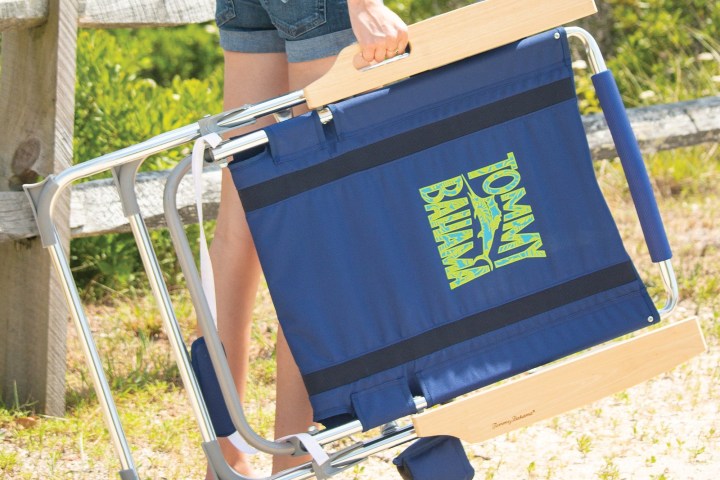 Person carries blue Tommy Bahama beach chair on sandy path.